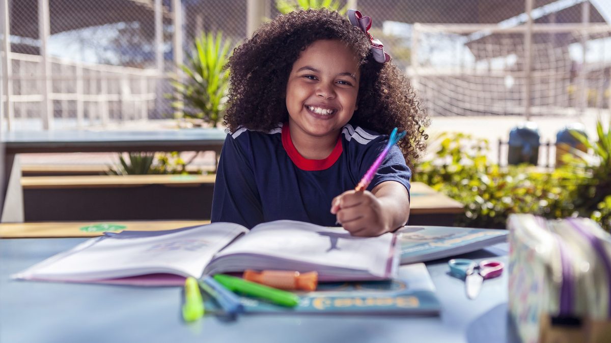 Uma criança feliz usando uniforme escolar, escrevendo com uma caneta de sereia em um livro, mostrando que o retorno à rotina escolar deve ser um momento feliz